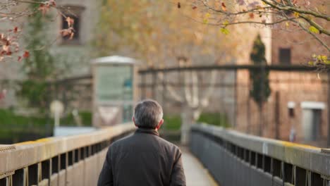 male adult with glasses and grey hair walks across a bridge away from the camera in an urban setting