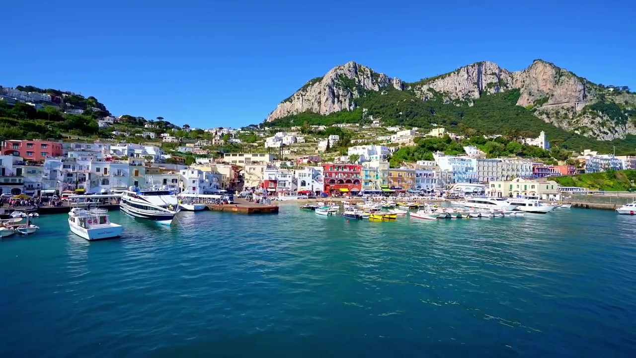 Ripresa con drone del porto di Marina Grande in Grecia con barche colorate ormeggiate lungo la banchina e case bianche che si ergono su una verde collina sotto la luce del giorno.