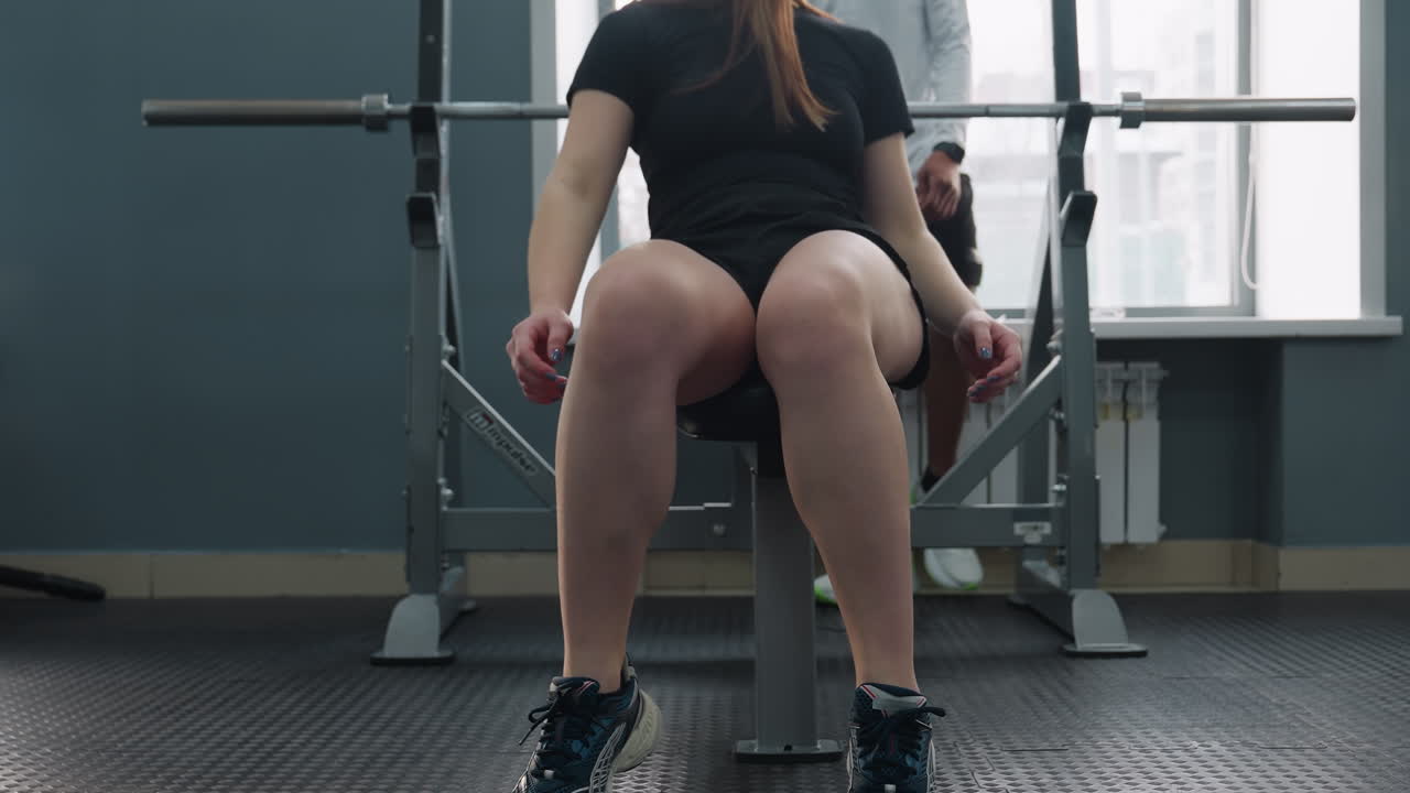 lower angle view of female client lying on bench pressing barbell with trainer approaching to provide spotting assistance under urban window light and modern gym equipment in background