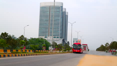 a red metro bus coming on the road, crossing the camera, low angle, close up, behind a tri building tower and couple of buildings, beautiful street lights and greenery on the road