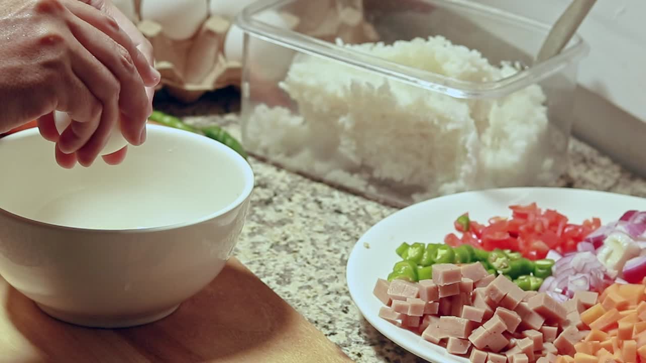Cracking eggs on a white bowl at a kitchen counter surrounded by leftover rice, diced vegetables and ham for fried rice recipe