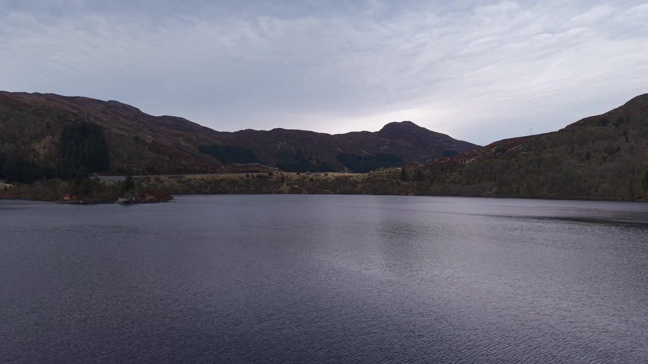 En rolig sø reflekterer de dæmpede farver fra en overskyet himmel, indrammet af bølgende bakker i Norge. Ideel til projekter, der fremhæver fredfyldte naturlandskaber