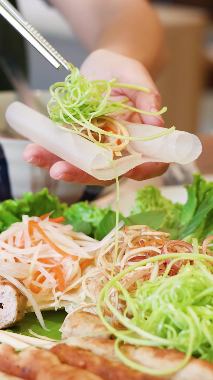 Bidikan close-up tangan merakit lumpia Vietnam tradisional dengan bumbu segar dan sayuran.