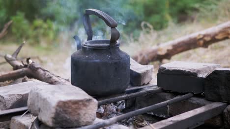 old black smoked kettle warms itself on grate of a smoking, fading bonfire. 4k