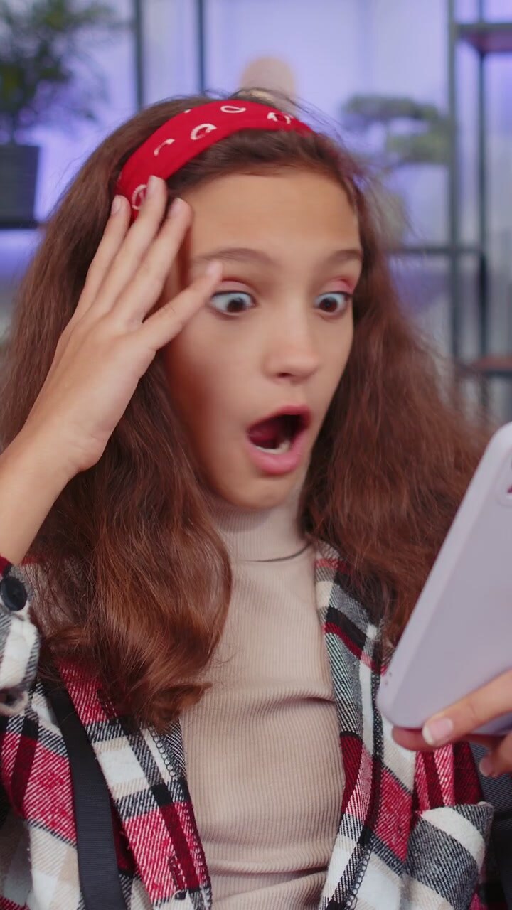 Bambina sul divano di casa che usa lo smartphone, digita e naviga, esclamando "wow, sì" dopo aver scoperto una grande vittoria.