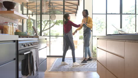 happy diverse couple in aprons dancing in sunny kitchen, copy space, slow motion