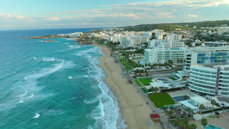 Bulliciosa-Ciudad-Costera-Aérea-De-Protaras-En-Chipre,-Vibrante-Tramo-De-Playa-De-Arena,-Olas-Espumosas-Del-Mar-Mediterráneo-Azul,-Hileras-De-Edificios-Modernos-Con-Un-Telón-De-Fondo-De-Terreno-Montañoso