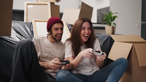 A-happy-and-engaged-couple,-a-brunette-girl-and-her-boyfriend-with-stubble-in-a-beige-T-shirt-play-video-games-using-two-joysticks-while-sitting-near-a-sofa-covered-with-a-plastic-cover-and-among-a-large-number-of-boxes-after-moving-to-a-new-apartment