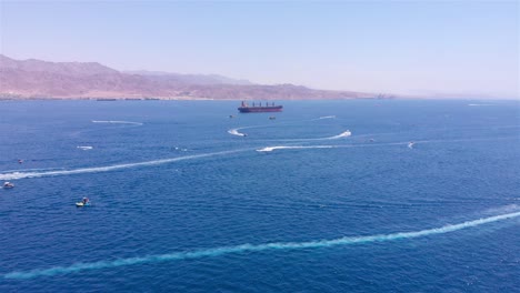 aerial view of a bay with jet skis, small boats, and a cargo ship against a mountainous backdrop