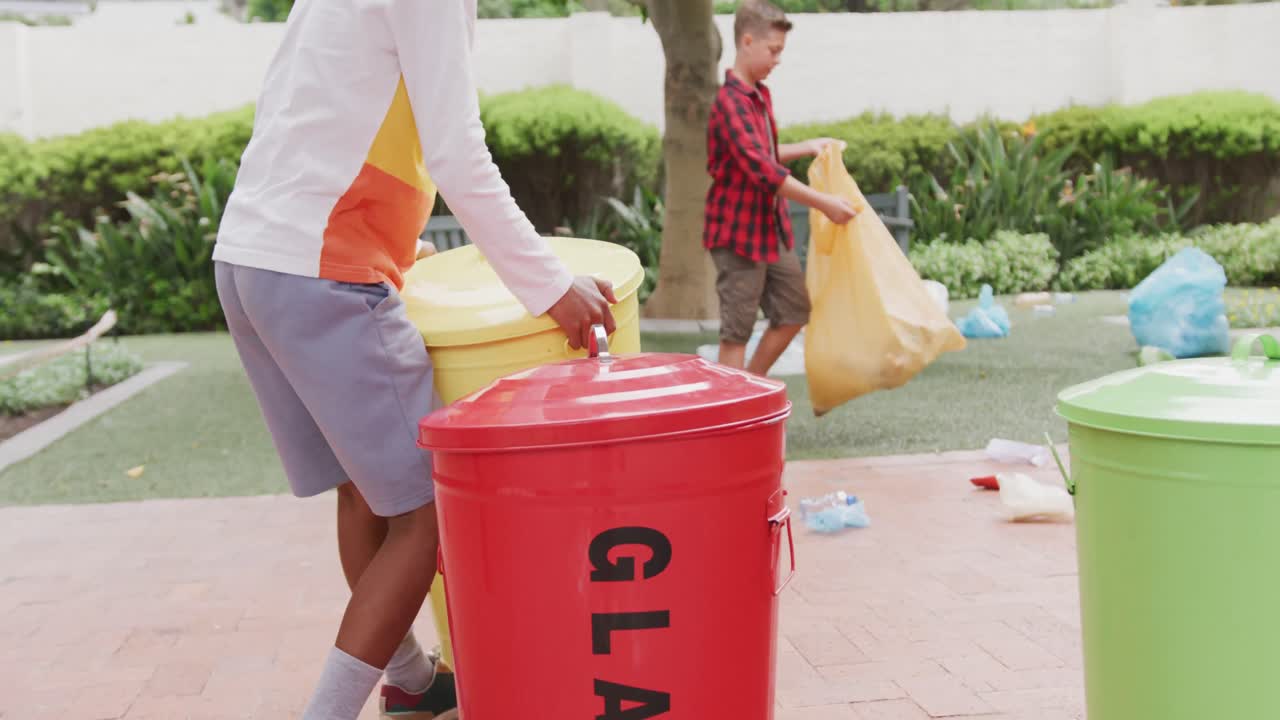 Video of two happy diverse schoolboys collecting rubbish for recycling in schoolyard