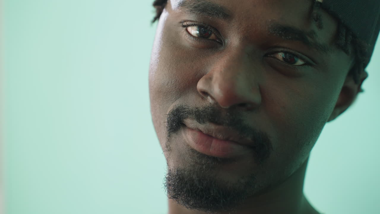 Close up of young man turning head toward camera after staring pensively, lost in thought, wearing cap backwards, showing contemplative expression on face against teal background in soft light