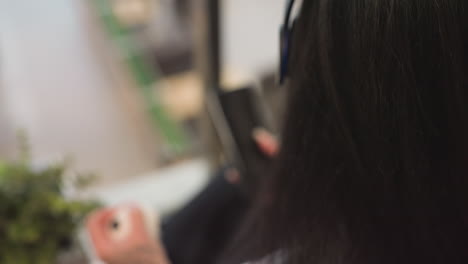 rear view of lady with long black hair wearing headphones holding phone and small ball in her hands, seated beside glass railing with floor below visible through transparent barrier in background