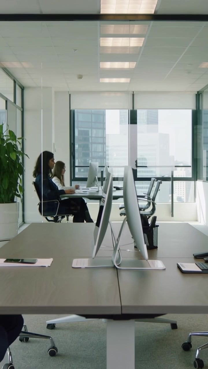 Video verticale: Due donne in blazer che digitano lavorando alla scrivania con monitor mentre un uomo passa davanti alle finestre.