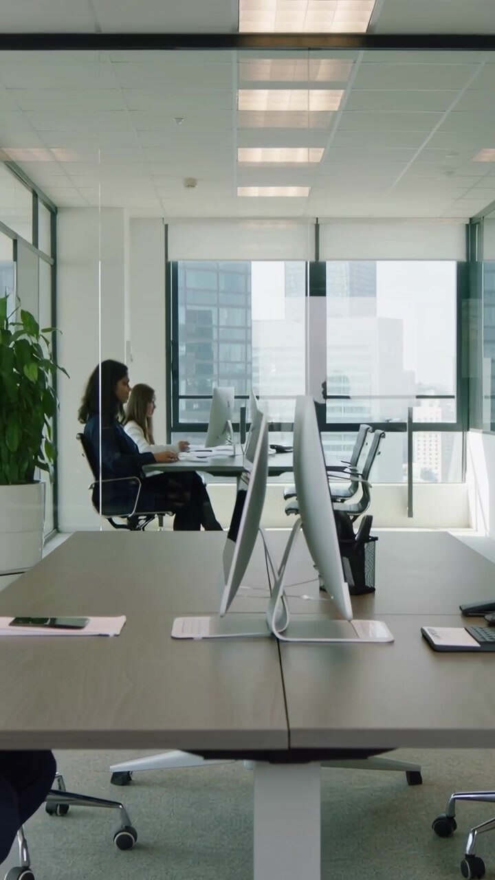 Video vertical: Dos mujeres con blazer escribiendo y trabajando en un escritorio con monitores mientras un hombre pasa por las ventanas.