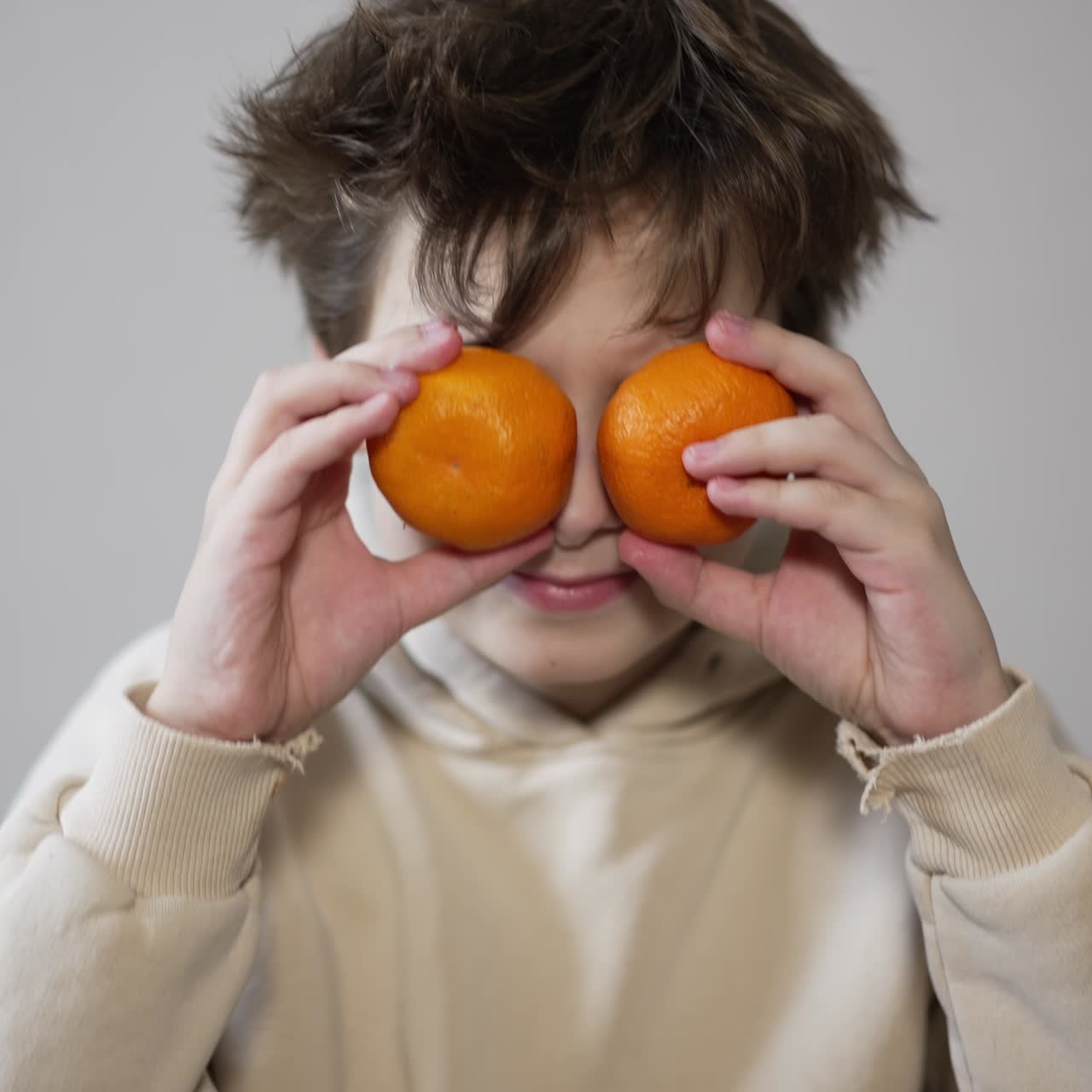 Handsome teenage kid sitting in front of camera. Kid puts some tangerines as his eyes. Close up