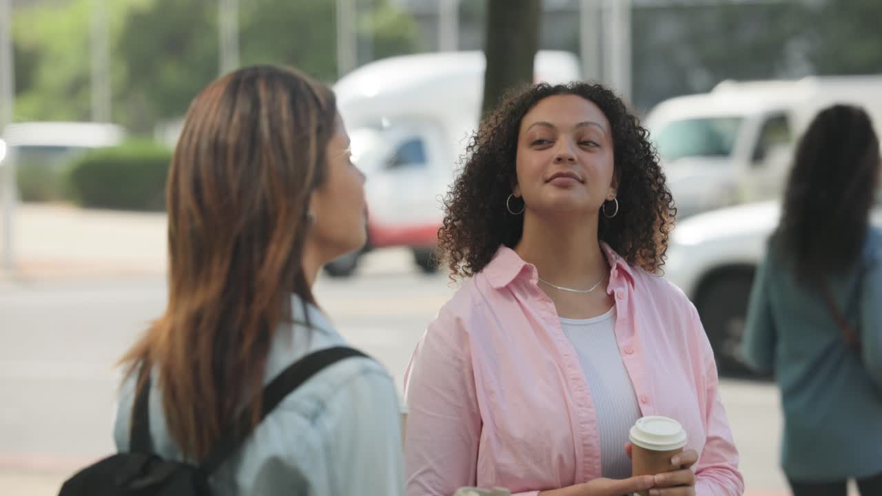 Mulheres diversas em pé na calçada com copos para viagem, a mulher de blusa rosa cheirando o café e verificando o telefone.
