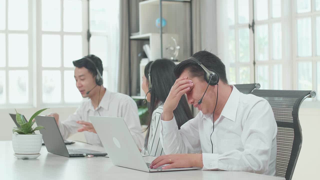 A Man Of Three Asian Call Center Agents Wearing Headset Is Tired Because Two Of His Colleagues Are Talking During Working At The Office