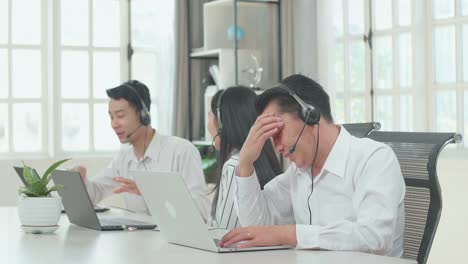 a man of three asian call center agents wearing headset is tired because two of his colleagues are talking during working at the office