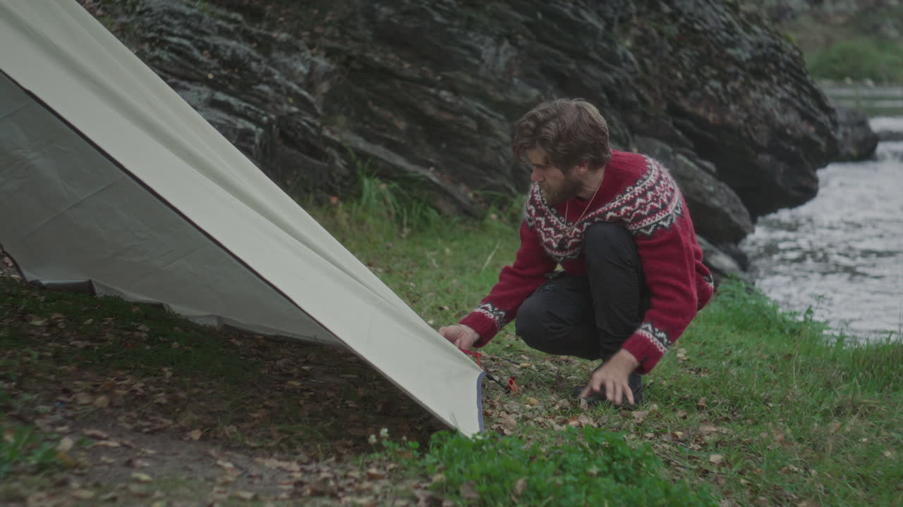Tourist Securing Tent Corner with Stone on Riverbank Campsite