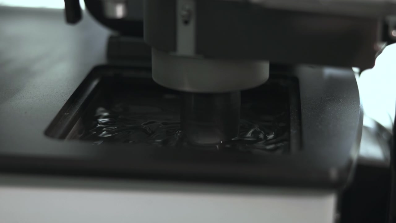 Low-angle close-up shows cylindrical probe or nozzle contacting shallow liquid inside rectangular tray within laboratory machine, creating ripples, small splashes, reflections, and cool rim lit