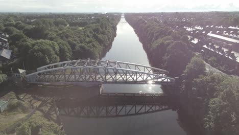 Vista-Aérea-Descendente-Hacia-El-Pintoresco-Puente-Peatonal-De-Tráfico-De-Arco-De-Acero-Antiguo-Sobre-El-Cruce-Del-Canal-De-Barcos-De-Manchester