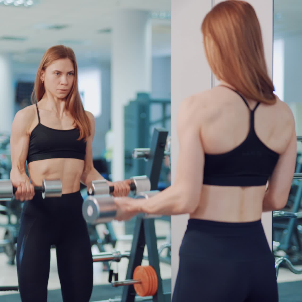 A beautiful woman in black leggings and top is doing an exercise for biceps using dumbbells opposite mirror in the gym. Blurred Background