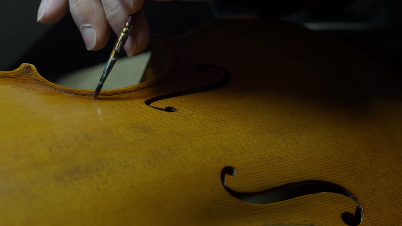 Luthier antiquing violin top plate near f-holes and purfling, applying pigment with brush and fingertip tampon to simulate aging, scratches, and authentic wear in a traditional finishing method
