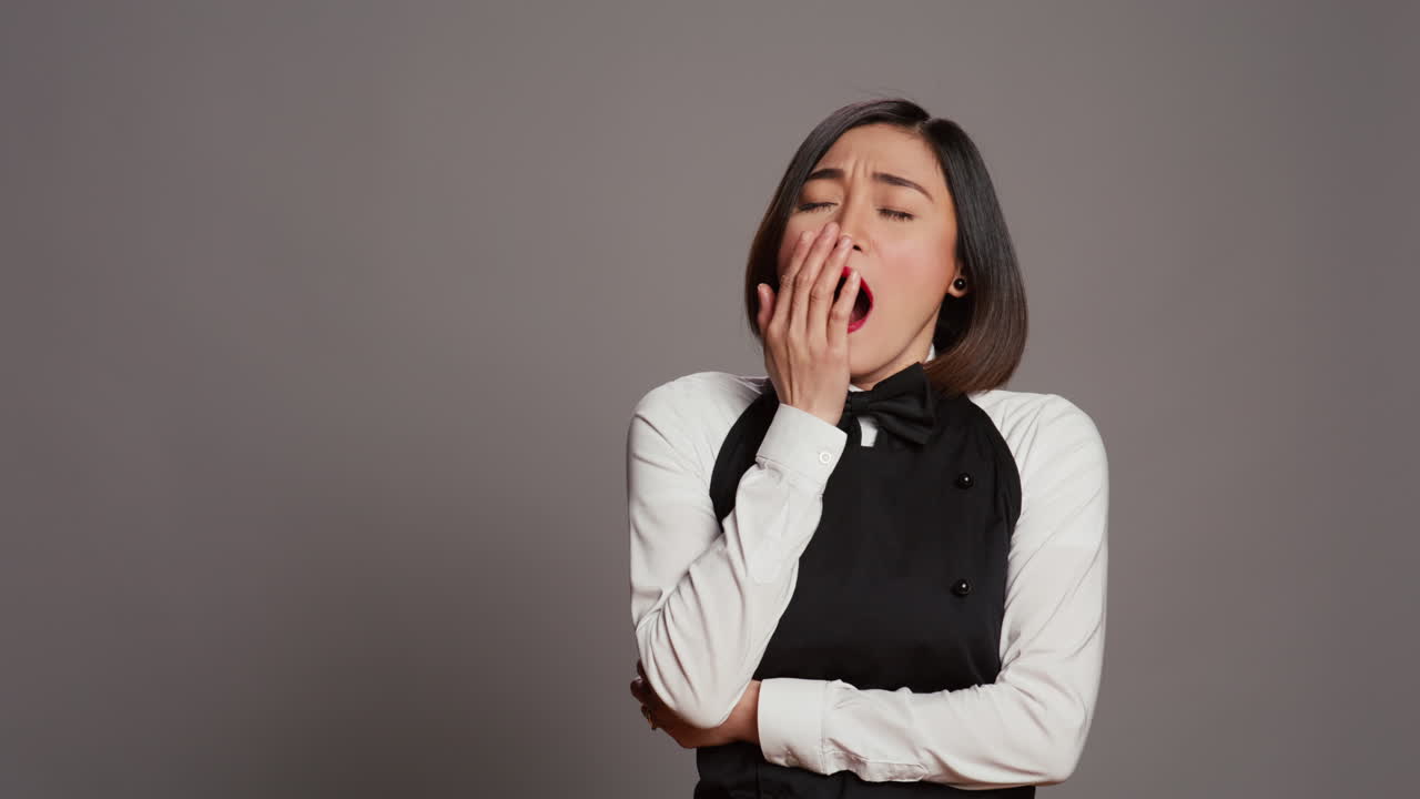 Exhausted waitress with apron yawning over grey background
