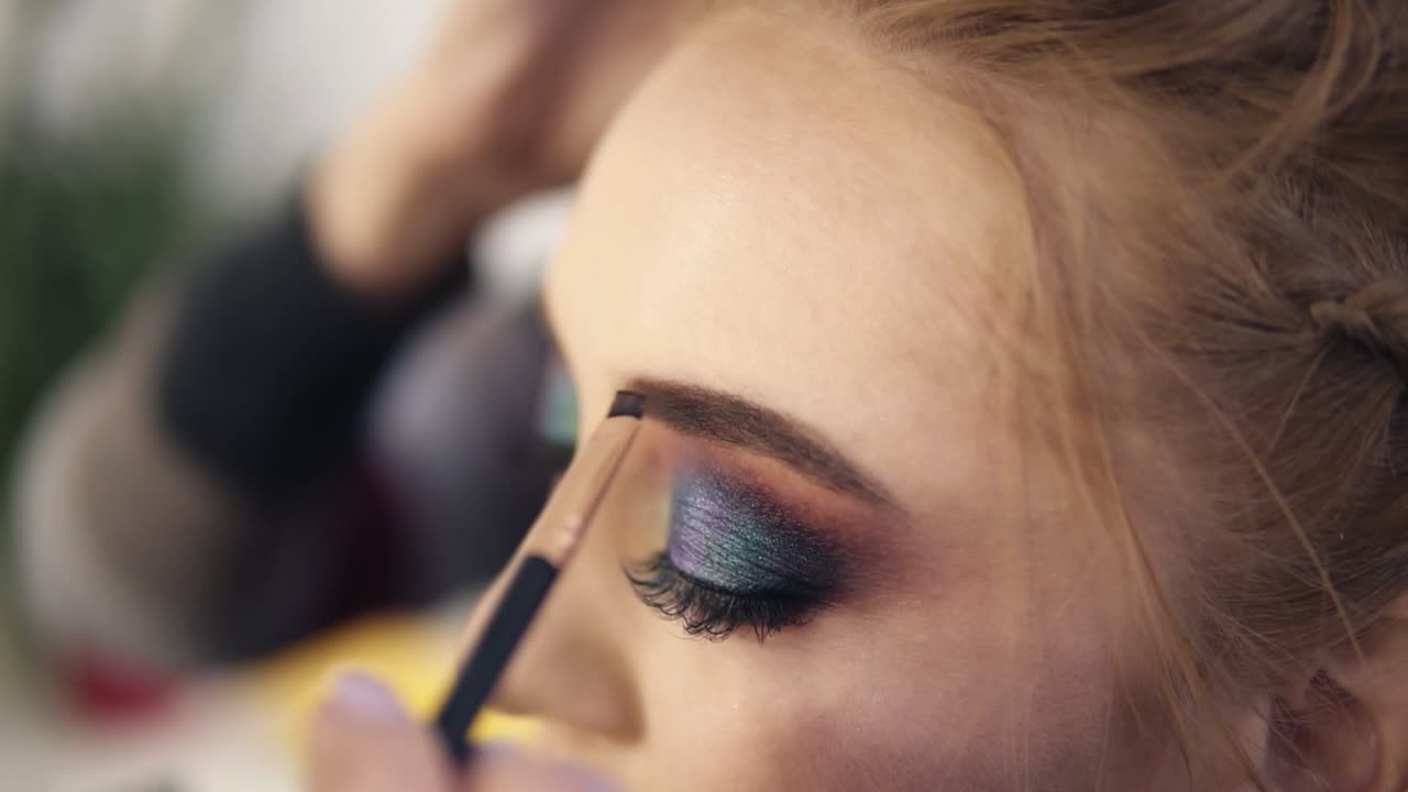 el estilista pone algunas sombras marrones usando un pincel y contorneando la ceja izquierda de una mujer bonita con ojos de humo. preparación. vista lateral
