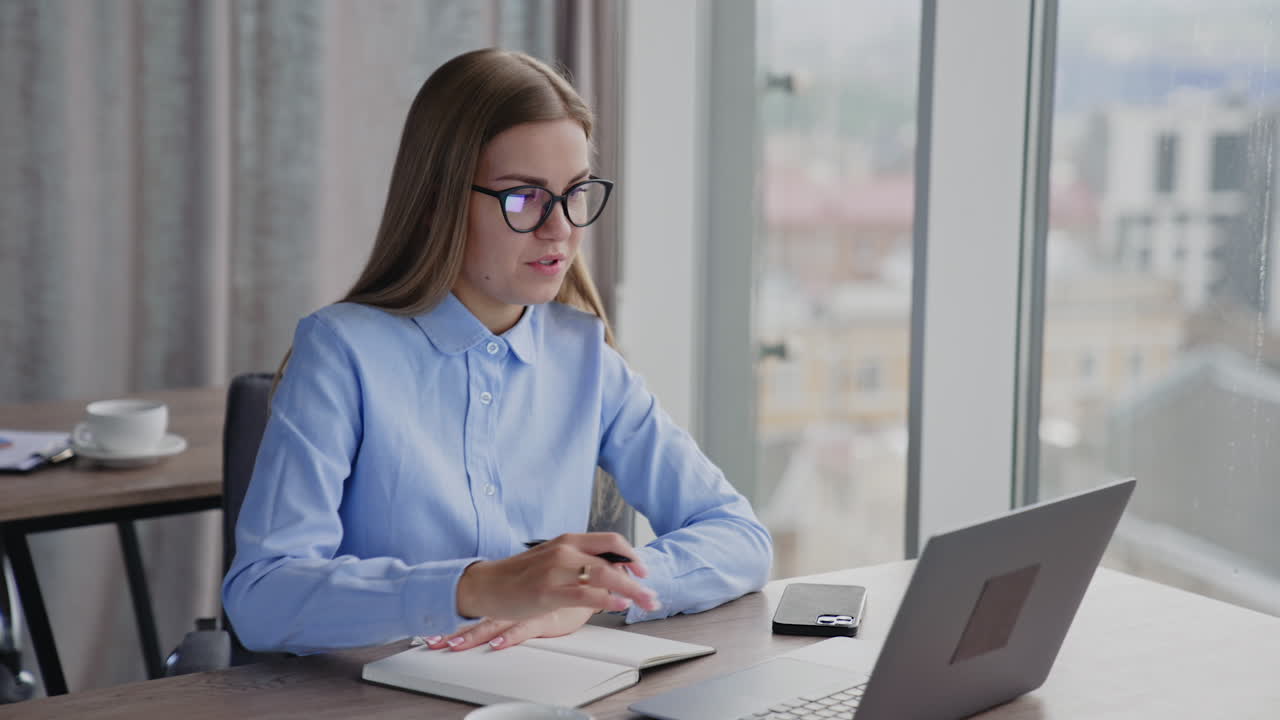 Female employee writing and talking. Woman looks at the screen of laptop and continues to write. Blurred backdrop.