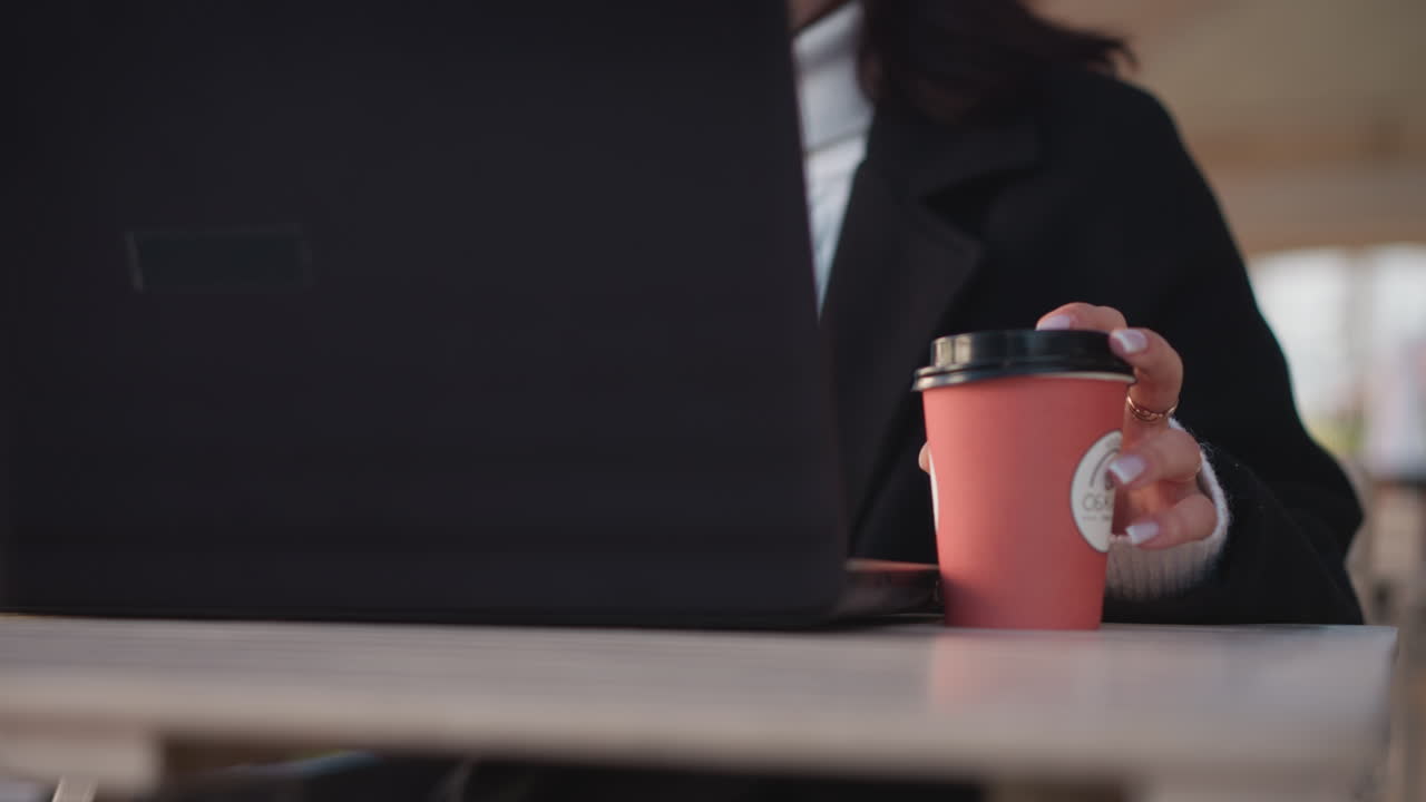 노트북에서 타이핑하는 동안 은 커피 컵을 부드럽게 들고 있는 여성의 손의 클로즈업, 멋진 매니큐어와 현대적인 작업 공간을 보여주는 야외 카페 또는 사무실의 흐릿한 배경