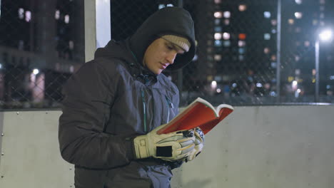 goalkeeper wearing hand glove in hoodie and beanie leaning against metallic goalpost reading book outdoors at night, illuminated by city lights