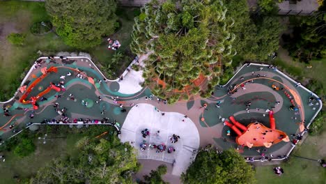 children’s playground with orange play structures, winding paths, and people gathered throughout, surrounded by trees near parque rivadavia in buenos aires. static top-down drone view