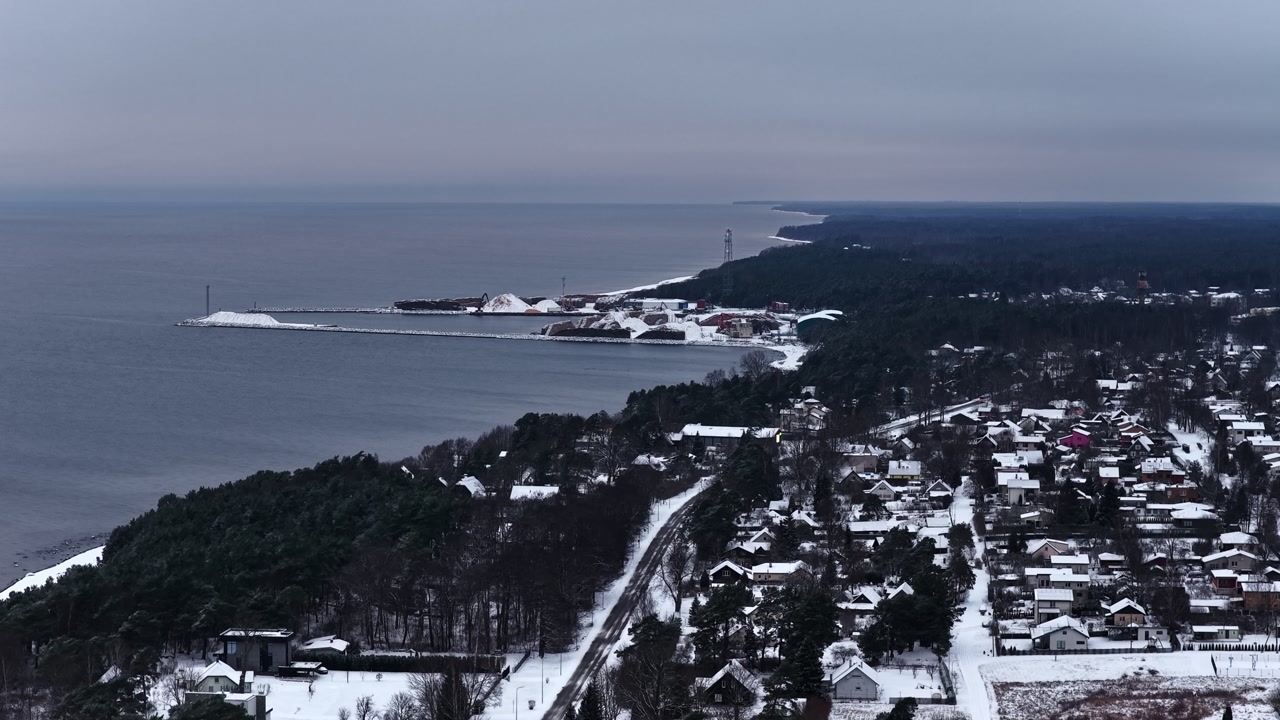 Drone aerial of a snowy coastal town along a long shoreline, with dark sea, pine forest, and winter rooftops under an overcast sky—wide Baltic coastline view.