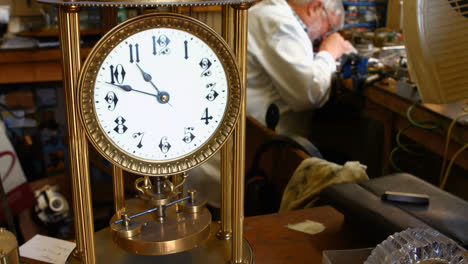 Vintage-Torsionspendeluhr-Auf-Dem-Tisch,-Während-Im-Hintergrund-Ein-Uhrmacher-Arbeitet