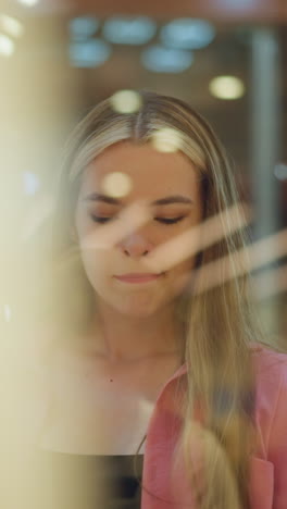 dama de ropa rosa mira a través del vidrio con una expresión contemplativa, reflejando luces borrosas y un letrero blanco brillante en el fondo, el vidrio refleja luces suaves y brillantes