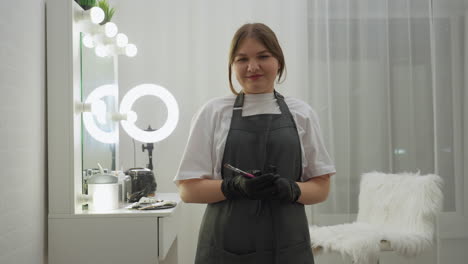 beautician in black gloves and apron stands in salon environment holding cosmetic brushes and looking at camera with calm expression. vanity mirror, ring light, and furry chair visible beside her