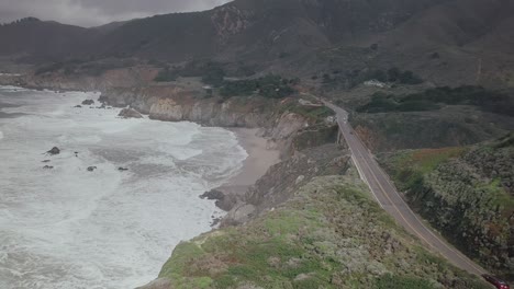 Descending-aerial-drone-shot-following-route-of-South-pacific-highway-along-rocky-terrain-of-coastal-route-as-waves-crash-in-against-side-of-cliff