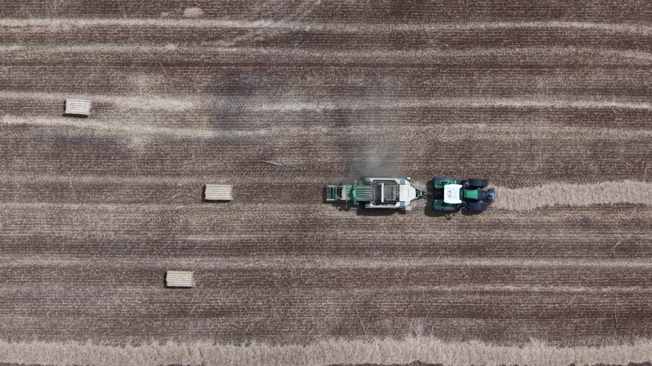 Una vista aerea cattura un trattore moderno e un'imballatrice che lavorano in un vasto campo raccolto, trasformando efficientemente la paglia in balle.