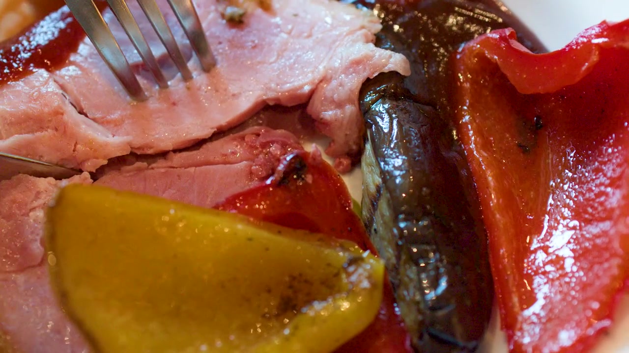 Un coltello e una forchetta tagliano il prosciutto arrosto servito con peperoni rossi e gialli grigliati.