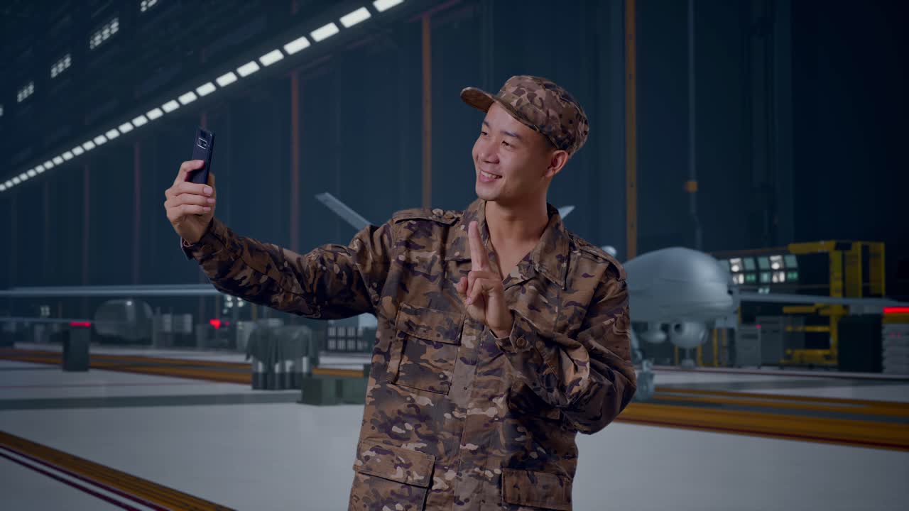Soldato asiatico usa lo smartphone per scattare una foto, in piedi con un moderno drone militare in un hangar di manutenzione.