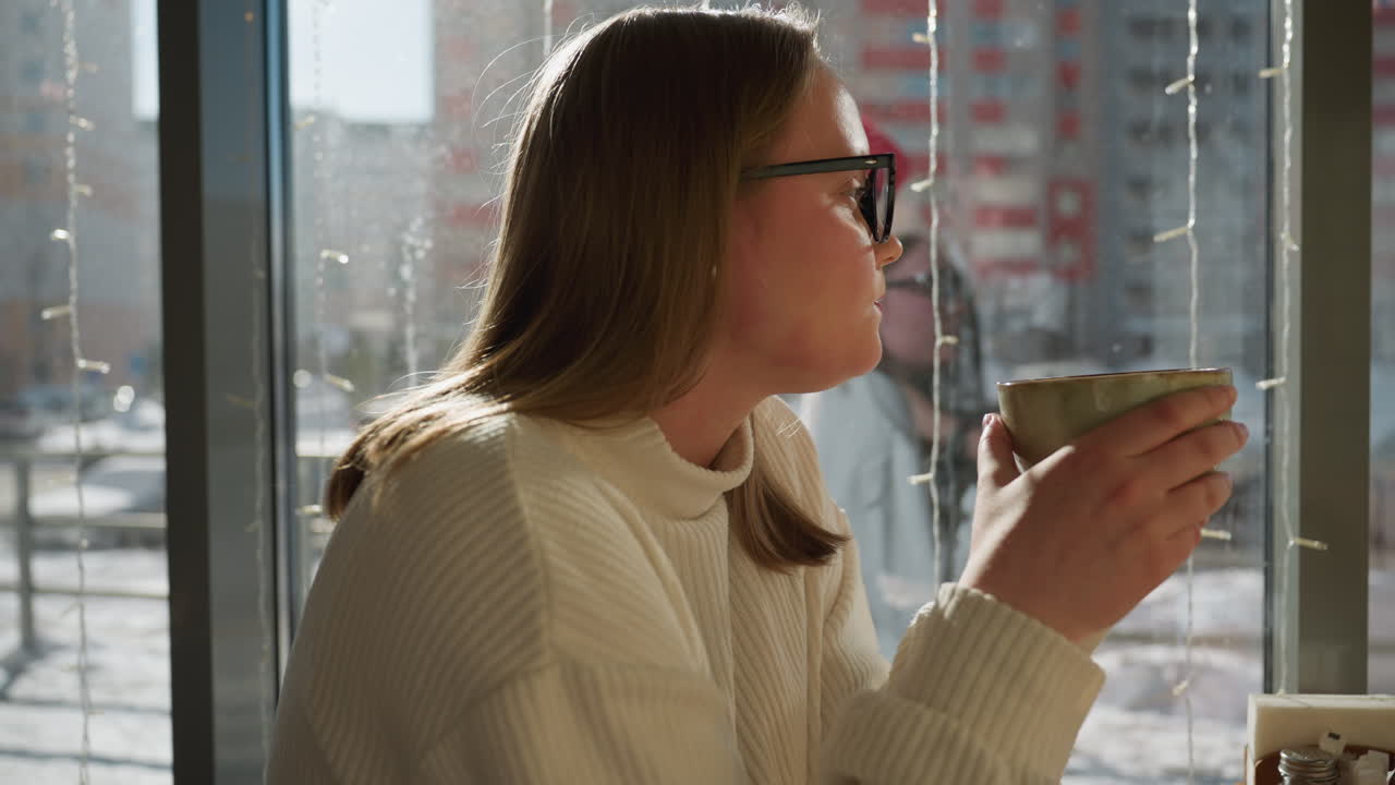 Freelancer sipping from cup while having breakfast inside cozy cafe, sunlight casting warm glow through glass window, snowy city street outside with parked cars and tall residential buildings visible