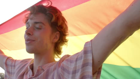 portrait of happy caucasian man holding rainbow lgbt flag on balcony at home, slow motion