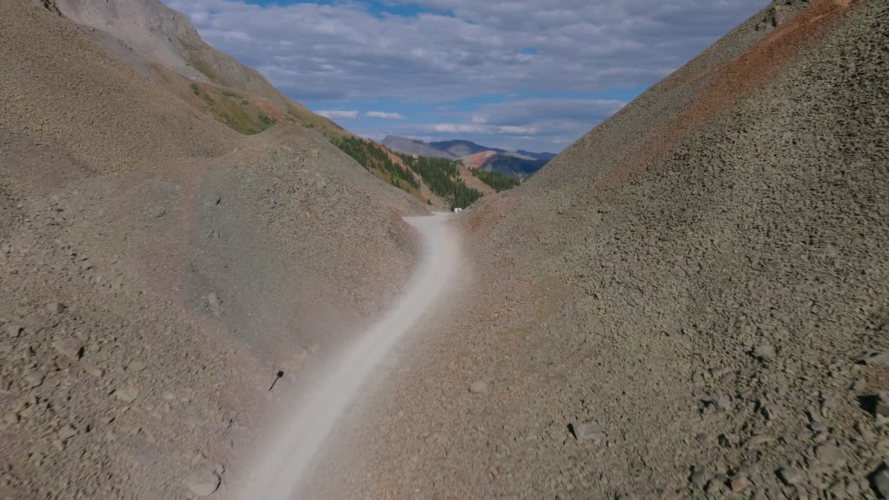 La strada di Ophir Pass si snoda ripidamente attraverso pendii montani instabili, strati sedimentari grezzi e la fragile geologia dell'alta regione di San Juan. Panoramica aerea in dolly su un SUV parcheggiato.