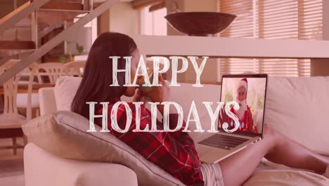 animation of happy holidays text over woman on laptop making christmas video call with santa claus