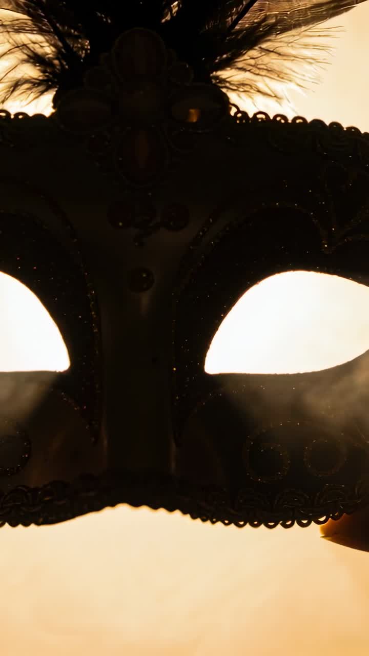 Video verticale: Una mano si estende a destra, sollevando una maschera nella calda luce dello studio, mostrando fumo e glitter.