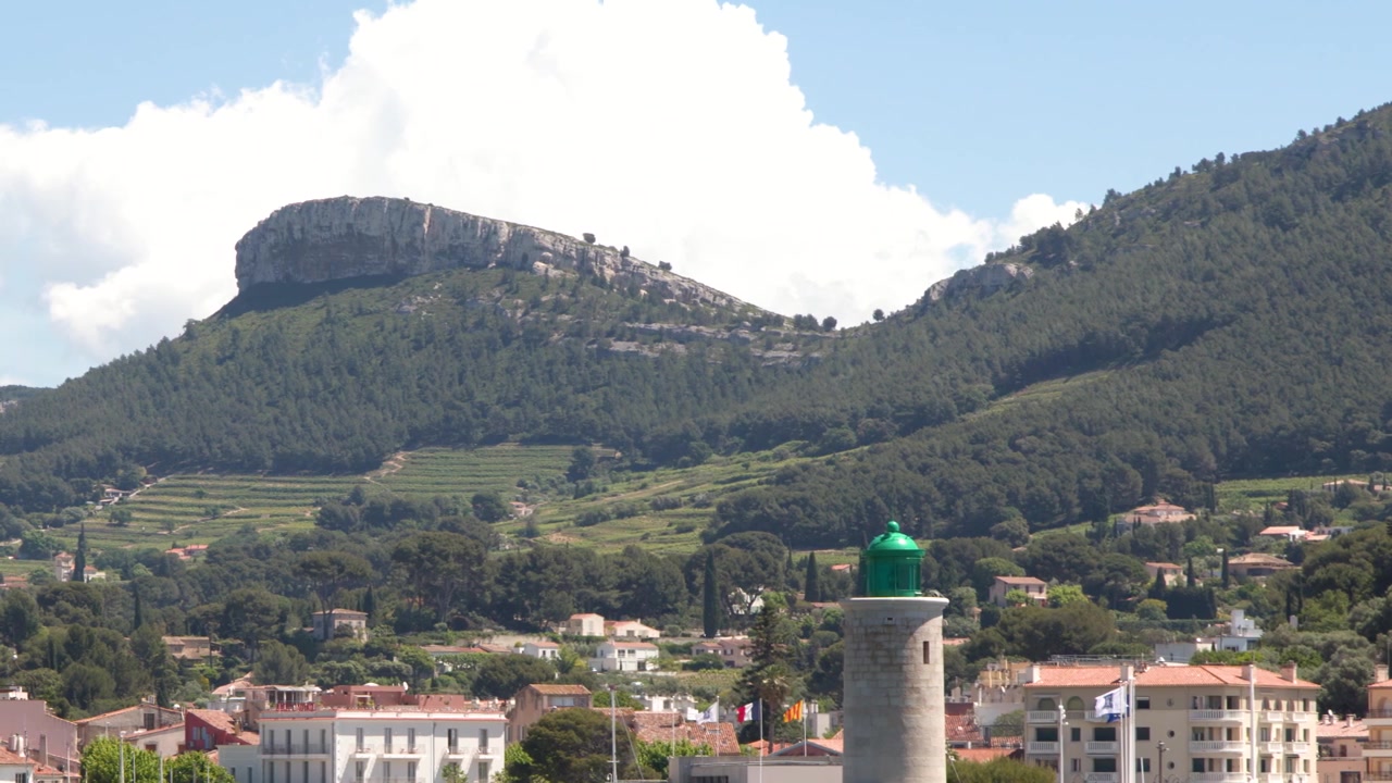 Ampia inquadratura statica di una città portuale mediterranea con un faro sullo sfondo di montagne lussureggianti.