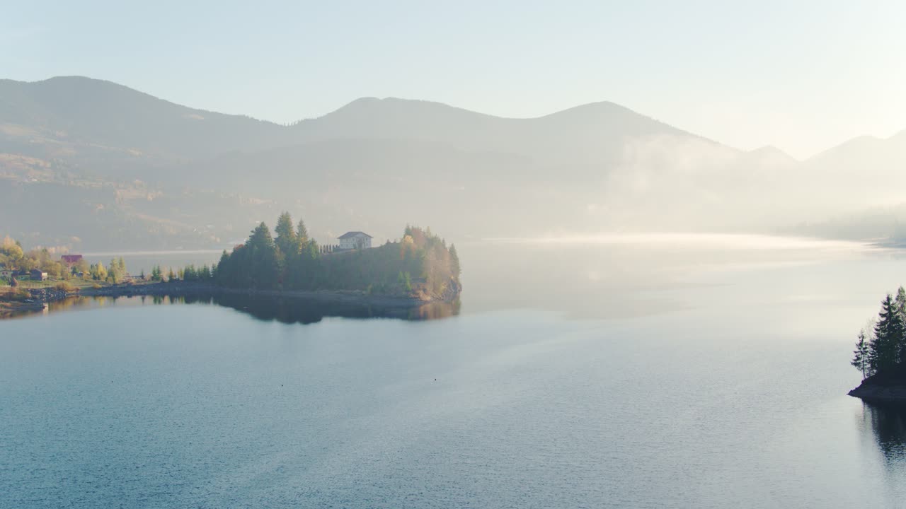 Aerial shot a lake and misty mountains at sunrise with house on island. Colibita, Romania