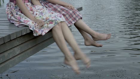 two children shake their bare feet above the water. they sit on a wooden bridge over the water next to each other.