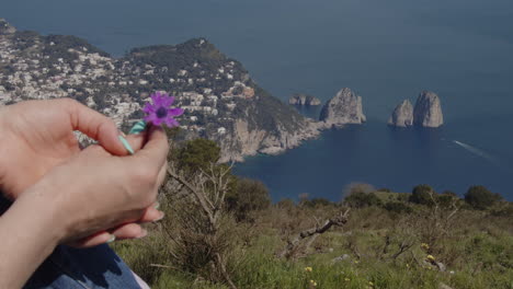 Girl-is-sitting-on-mountain,-holding-a-flower,-looking-at-faraglioni-Capri,-italy